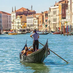 A gondola with a gondolier and passengers navigates a canal in Venice, surrounded by colorful historic buildings and boats on the Grand Canal.
