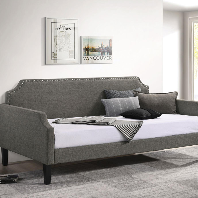 A gray upholstered daybed with nailhead trim features a curving backrest, bolster and square pillows, set against a wall with framed city posters.
