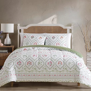 A bed features a patterned comforter and pillow set with floral designs against a neutral background. The space is styled with a wicker headboard, a wooden side table with a lamp, and decorative branches in a vase.