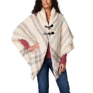 A woman wears a cozy, plaid toggle wrap with a fringed hem from St. John's Bay, featuring a shawl collar, toggle closures, and neutral tones, paired with a long-sleeve top and blue jeans.