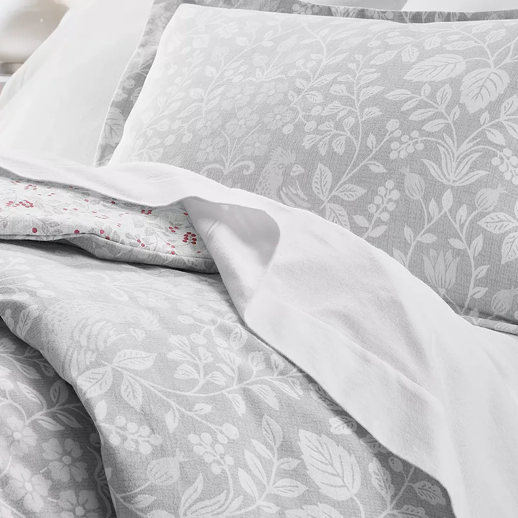 A set of bedding featuring a light gray and white floral pattern with decorative leaves and berries, paired with plain white sheets for contrast.