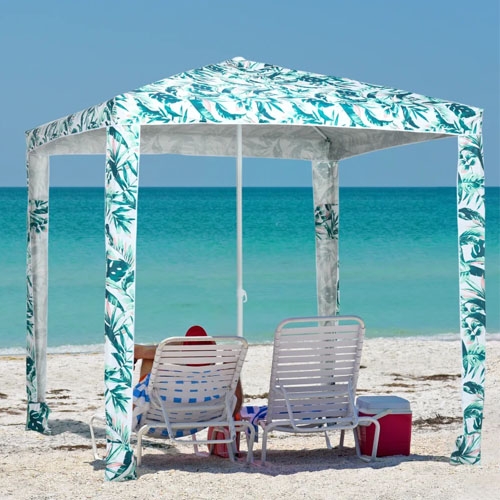 A tropical-patterned canopy with two folding chairs and a cooler on a sandy beach.