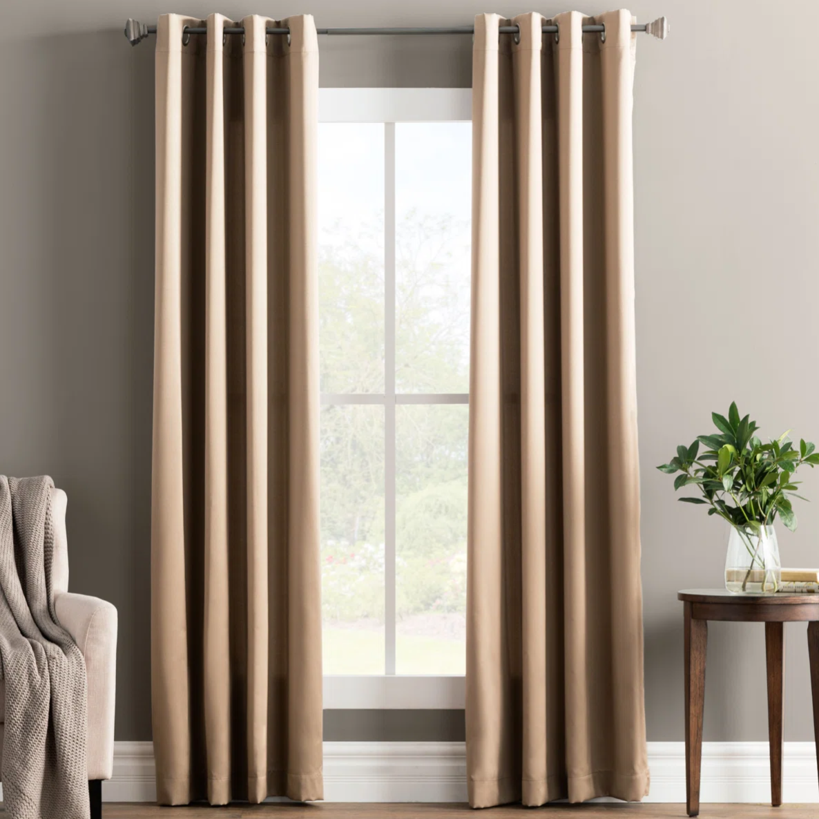 Tan curtains hanging on a rod with a modern grommet design, next to a beige sofa with a knitted throw. A small wooden table holds a vase with greenery.