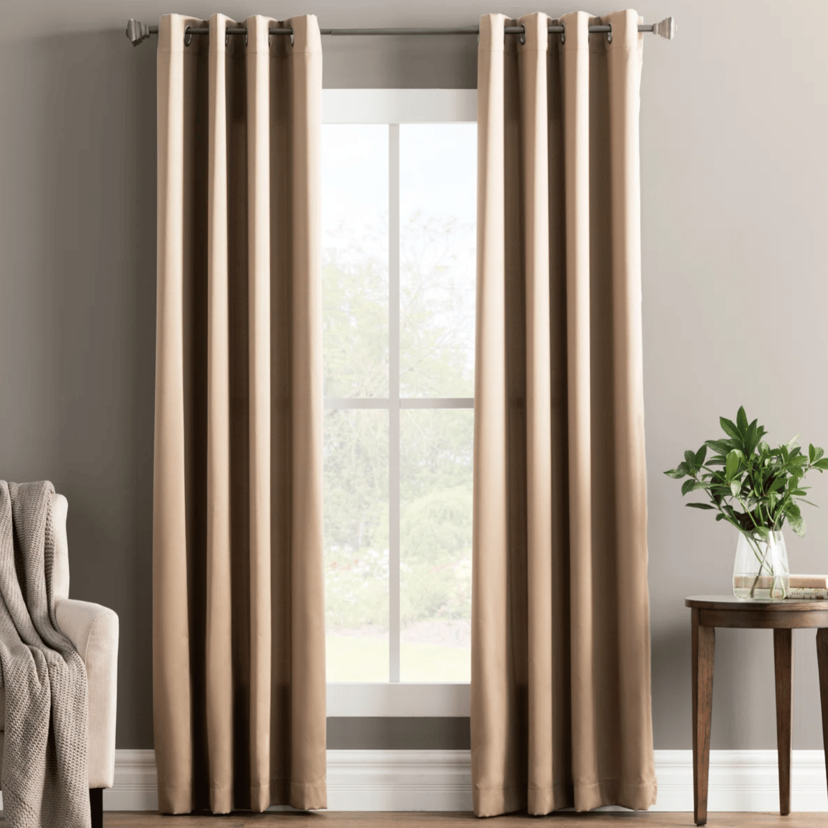 Tan curtains hanging on a rod with a modern grommet design, next to a beige sofa with a knitted throw. A small wooden table holds a vase with greenery.