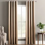 Tan curtains hanging on a rod with a modern grommet design, next to a beige sofa with a knitted throw. A small wooden table holds a vase with greenery.