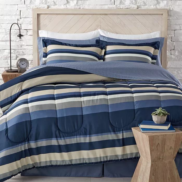 A bed is dressed with a striped blue, beige, and white comforter set, including matching pillows and a blue bed skirt. The style is complemented by a wooden headboard, a rustic side table with books and a small plant, and white brick wall decor.