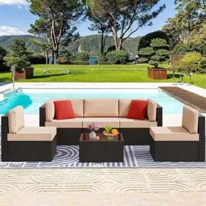 Outdoor wicker patio furniture set with beige cushions and red pillows, placed by a pool with a garden and trees in the background. The set includes a sofa, chairs, and a glass-top coffee table with fruit and a flower vase.