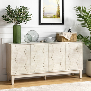 A white sideboard with a geometric pattern is adorned with a green vase holding leafy branches, glass plates, and a wicker basket with wine. A painting hangs above, and a potted plant stands nearby, set against a white wall and light wood floor.