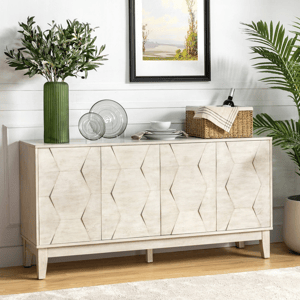A white sideboard with a geometric pattern is adorned with a green vase holding leafy branches, glass plates, and a wicker basket with wine. A painting hangs above, and a potted plant stands nearby, set against a white wall and light wood floor.