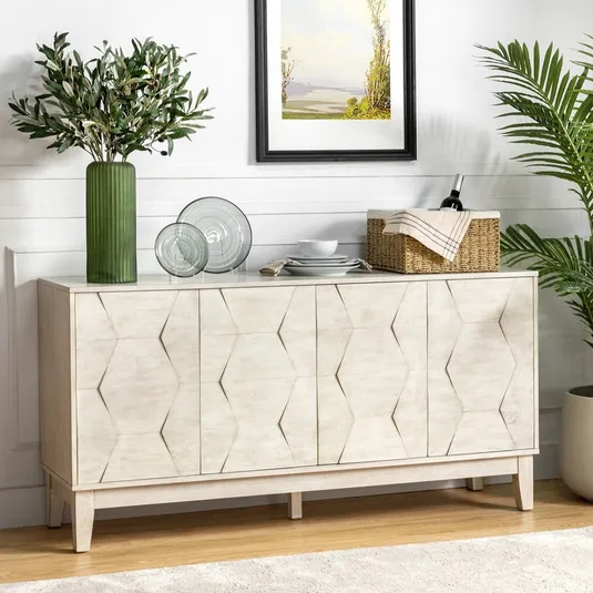 A white sideboard with a geometric pattern is adorned with a green vase holding leafy branches, glass plates, and a wicker basket with wine. A painting hangs above, and a potted plant stands nearby, set against a white wall and light wood floor.