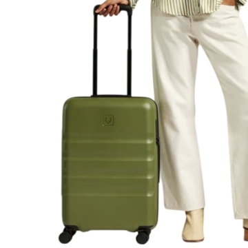 A person stands next to a green hard-shell carry-on suitcase with four wheels and an extendable handle, wearing white pants, a striped shirt, and beige boots.