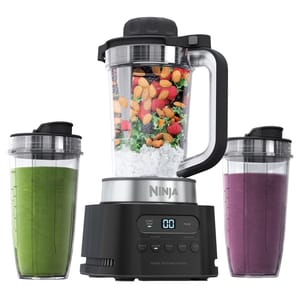A Ninja Blender with a sleek black and silver design includes a main pitcher filled with fruits, nuts, and ice, accompanied by two personal blending cups with green and purple smoothies.