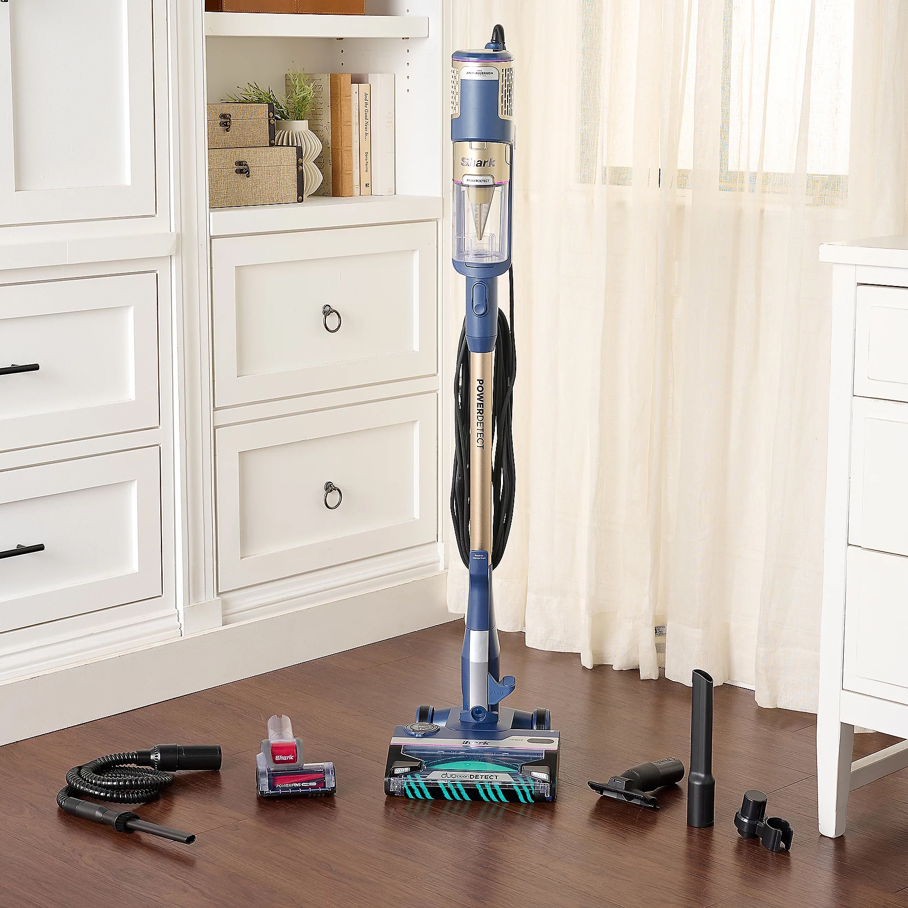 A blue Shark vacuum cleaner with a detachable dust canister is shown alongside several cleaning attachments and tools, set in a room with white cabinetry and light wooden flooring.