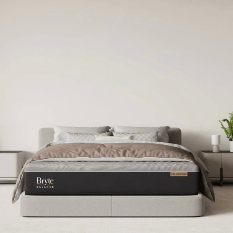 A modern bedroom setup featuring a Bryte Balance mattress with a \“Pro Conform\“ label. The room is decorated with neutral bedding, two bedside tables, a plant, and a small clock.