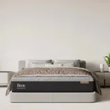 A modern bedroom setup featuring a Bryte Balance mattress with a \“Pro Conform\“ label. The room is decorated with neutral bedding, two bedside tables, a plant, and a small clock.