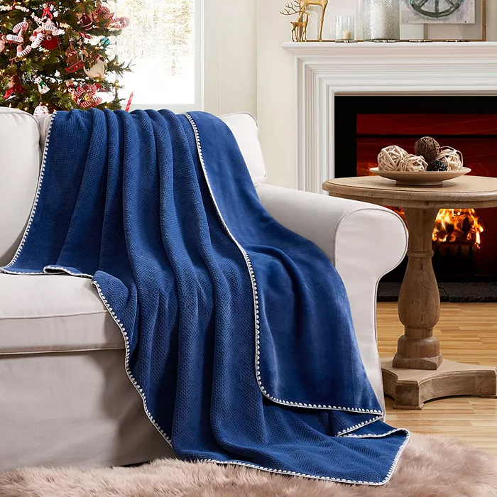 A blue Evergreen Ivy Embossed Waffled Throw draped over a beige sofa with white trim, in a cozy living room featuring a decorated Christmas tree and a fireplace.