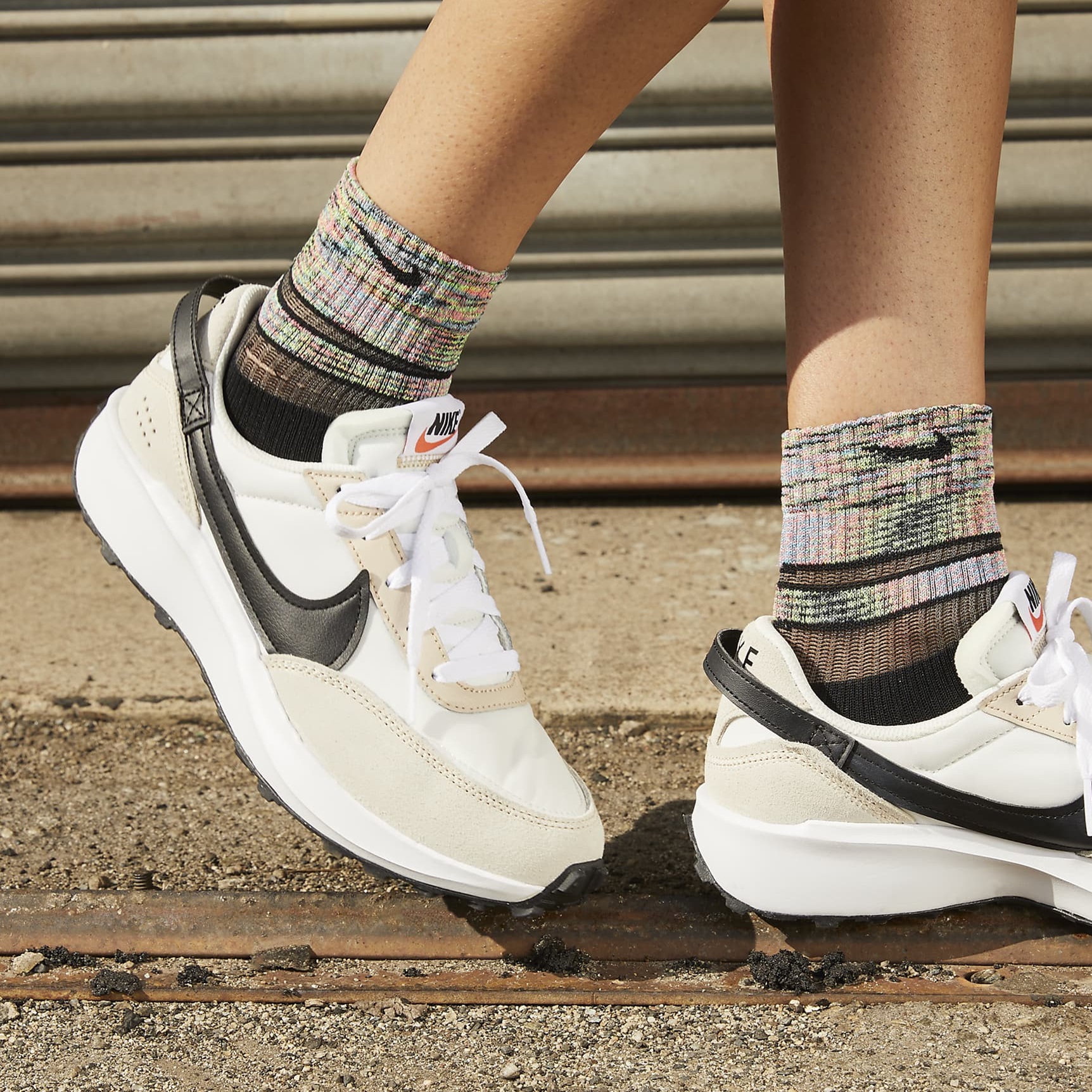 A pair of Nike sneakers with multi-colored socks worn by someone standing on a gritty surface.