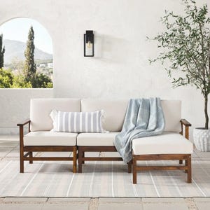 Outdoor wooden sofa set with beige cushions, striped pillow, and a blue plaid throw, situated on a light striped area rug. Features a wall-mounted lantern and potted plant.