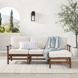 Outdoor wooden sofa set with beige cushions, striped pillow, and a blue plaid throw, situated on a light striped area rug. Features a wall-mounted lantern and potted plant.