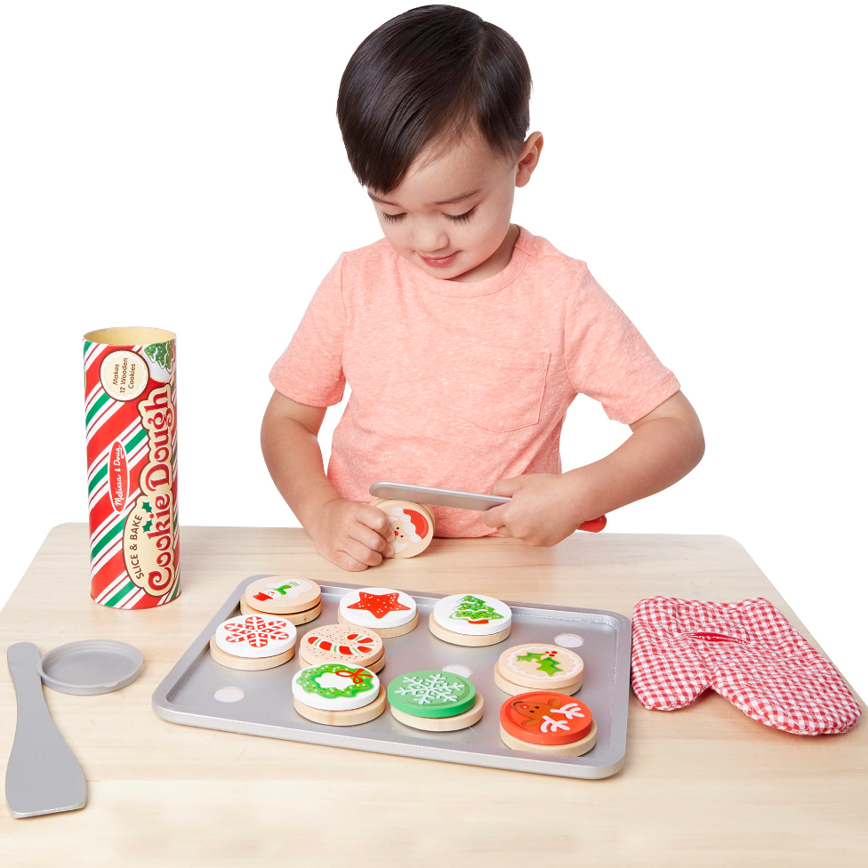 A child playing with a toy baking set, including a rolling pin, decorated cookies, a baking tray, an oven mitt, and a cookie dough tube.