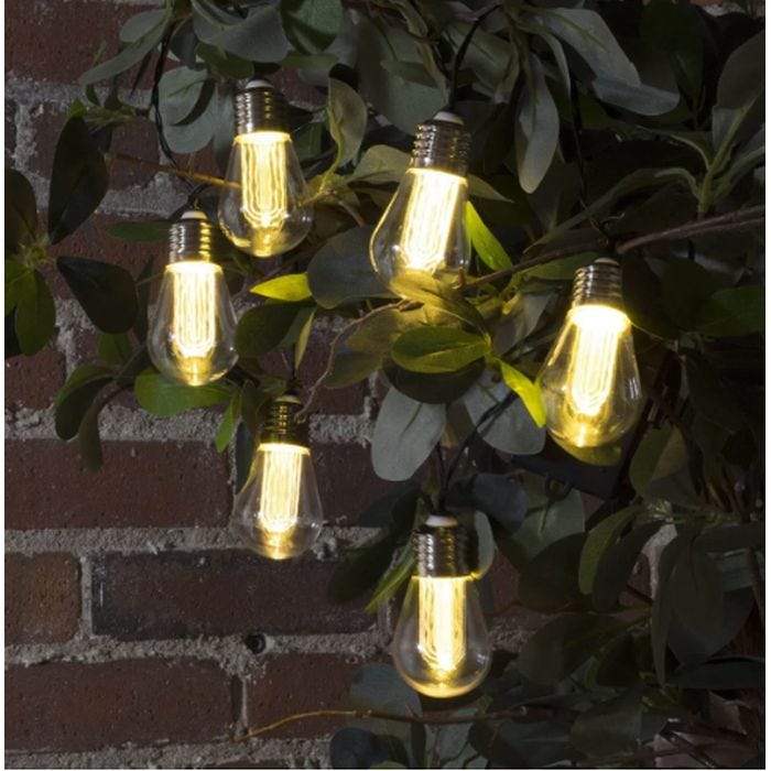 String lights designed to resemble vintage-style light bulbs are hung among leafy branches against a brick wall, creating a warm, decorative ambiance.