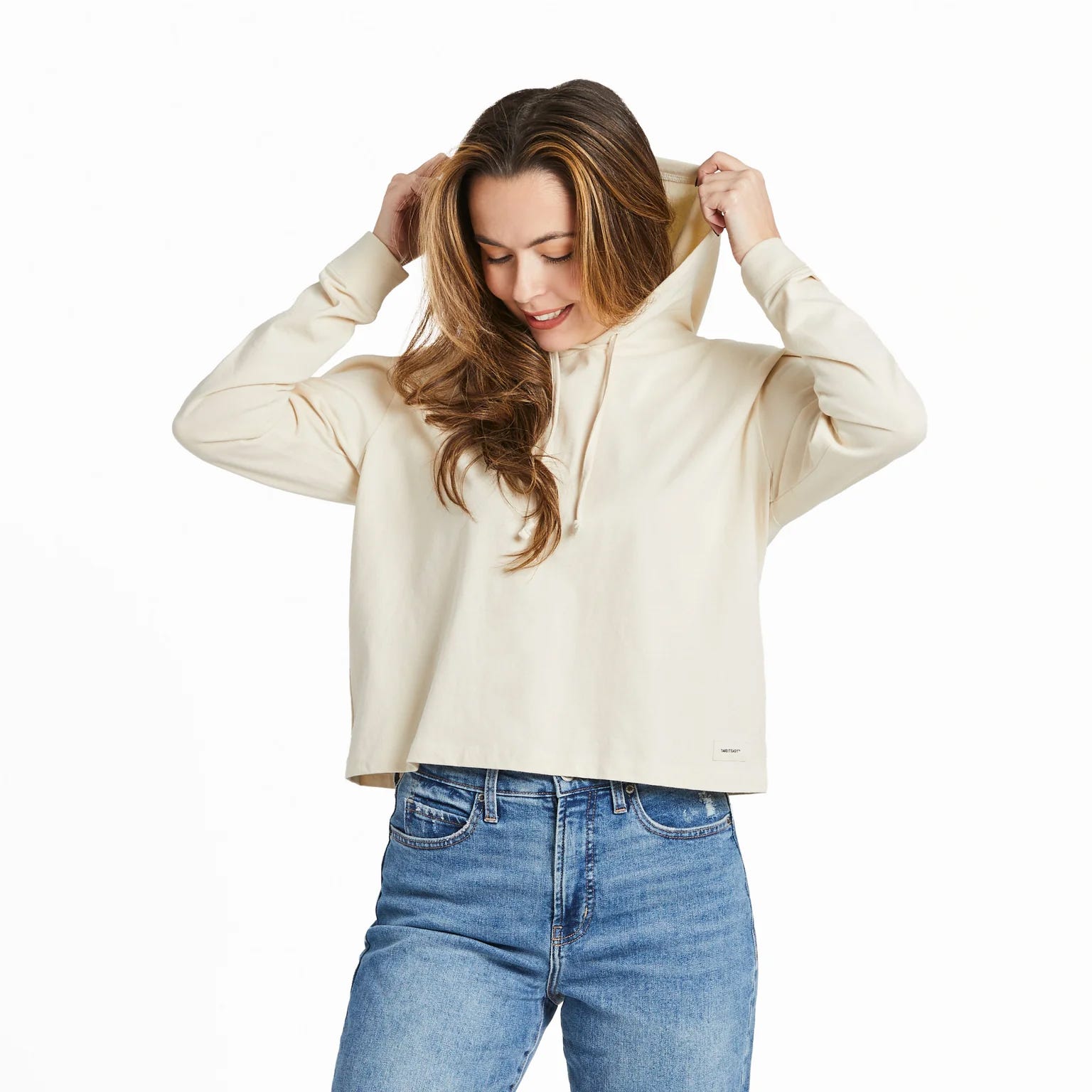 A woman wearing a cream-colored hoodie and blue jeans, with her hands adjusting the hood.