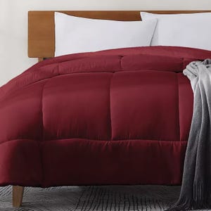A deep red comforter is displayed on a bed with a wooden headboard, with white pillows and a gray blanket draped over one side.
