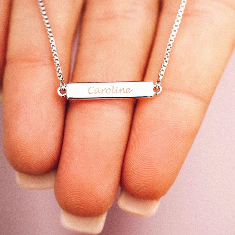 A silver bar bracelet features a delicate chain and a rectangular bar engraved with the name \“Caroline.\“ The design is simple and elegant.