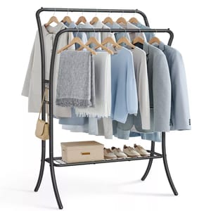 A double-rod clothes rack featuring two tiers for hanging clothes, includes wooden hangers with a mix of coats, shirts, and scarves. The lower shelf holds a box and shoes, and a small beige bag hangs on the side. The frame is dark metal.