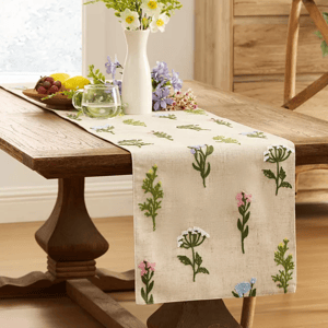 A wooden table is decorated with a floral embroidered table runner and a white vase of flowers. A small clear mug and a plate of fruit are also placed on the table.