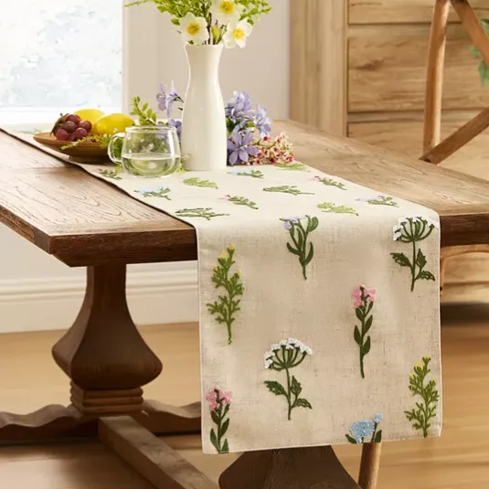 A wooden table is decorated with a floral embroidered table runner and a white vase of flowers. A small clear mug and a plate of fruit are also placed on the table.