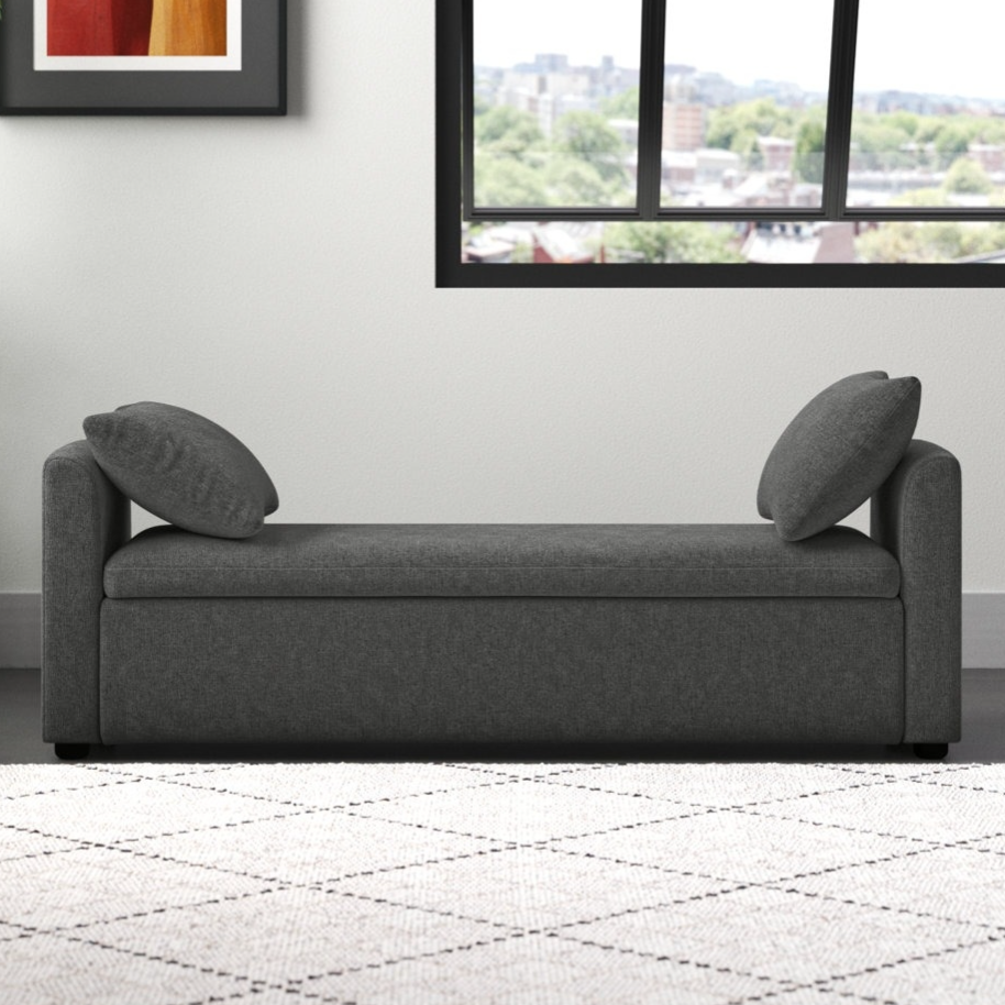 A gray upholstered chaise lounge with two cushions is set against a white wall. There's a framed abstract art piece and a large window, along with a potted plant on the left and a light-colored patterned rug on the floor.