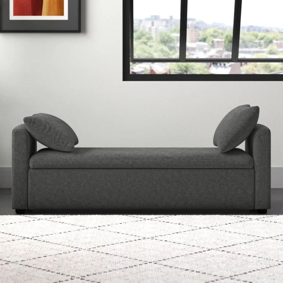 A gray upholstered chaise lounge with two cushions is set against a white wall. There's a framed abstract art piece and a large window, along with a potted plant on the left and a light-colored patterned rug on the floor.