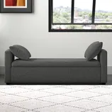 A gray upholstered chaise lounge with two cushions is set against a white wall. There's a framed abstract art piece and a large window, along with a potted plant on the left and a light-colored patterned rug on the floor.