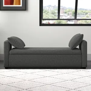 A gray upholstered chaise lounge with two cushions is set against a white wall. There's a framed abstract art piece and a large window, along with a potted plant on the left and a light-colored patterned rug on the floor.