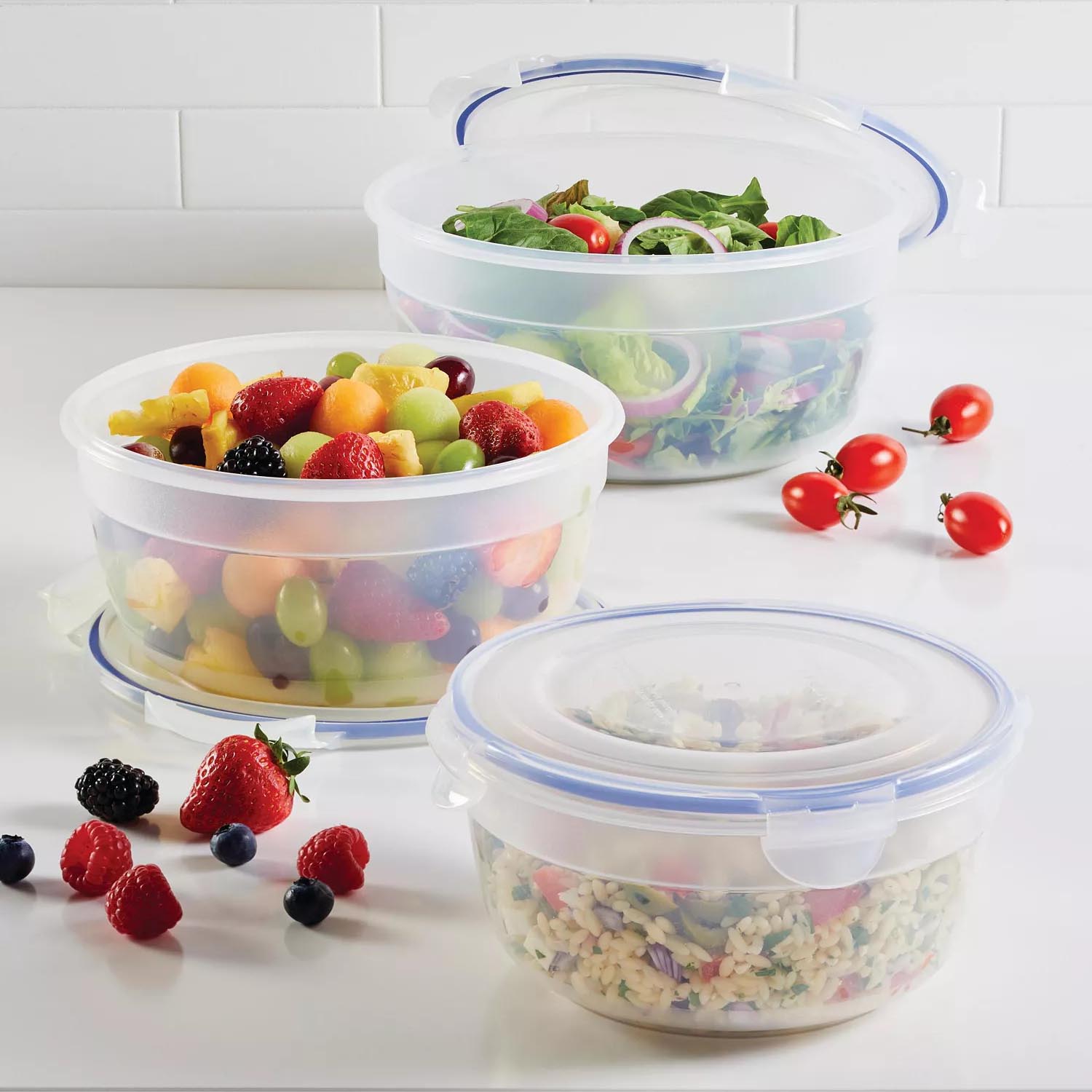 Three round, clear plastic food storage containers with blue sealing lids are shown, containing assorted fruits, salad, and a pasta dish. Scattered around are cherry tomatoes and various berries.