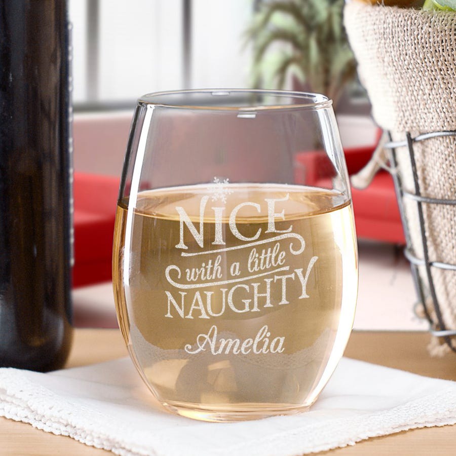 A stemless wine glass with the text \“NICE with a little NAUGHTY\“ and the name \“Amelia\“ etched on it, partially filled with white wine, placed on a white napkin.