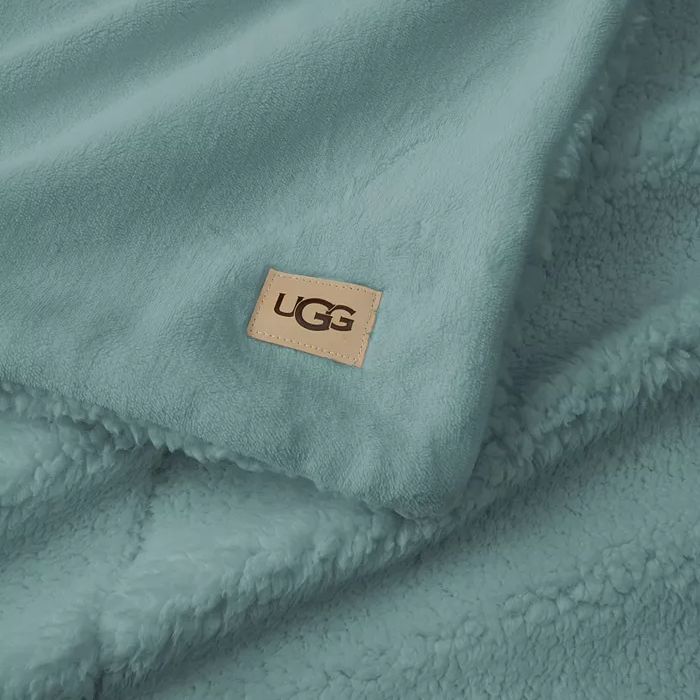 A soft blue UGG Cozy Throw featuring a plush, fleece texture with a visible UGG logo patch. Ideal for warmth and comfort.