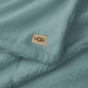 A soft blue UGG Cozy Throw featuring a plush, fleece texture with a visible UGG logo patch. Ideal for warmth and comfort.