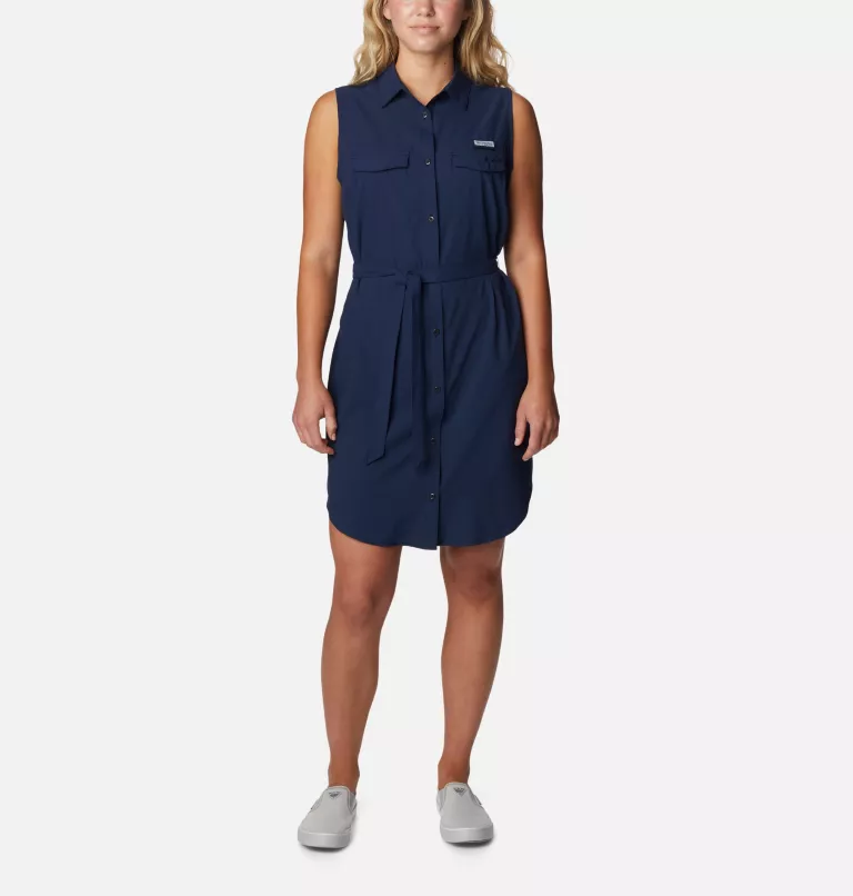 A woman models a sleeveless navy blue shirt dress with a collar, chest pockets, and a tied waist. She's wearing white slip-on shoes.