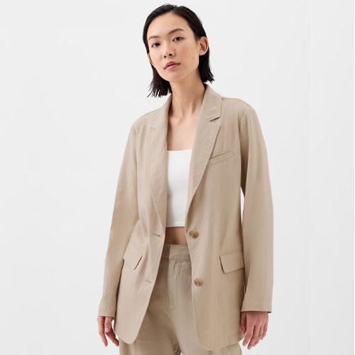 A woman models a beige linen-blend blazer with a relaxed fit, notched lapels, and front button closure, paired with a white top.