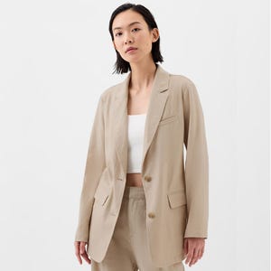 A woman models a beige linen-blend blazer with a relaxed fit, notched lapels, and front button closure, paired with a white top.
