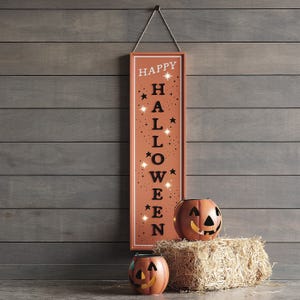A vertical \“Happy Halloween\“ sign hangs against a wooden wall, accompanied by two decorative pumpkin lanterns on a hay bale.