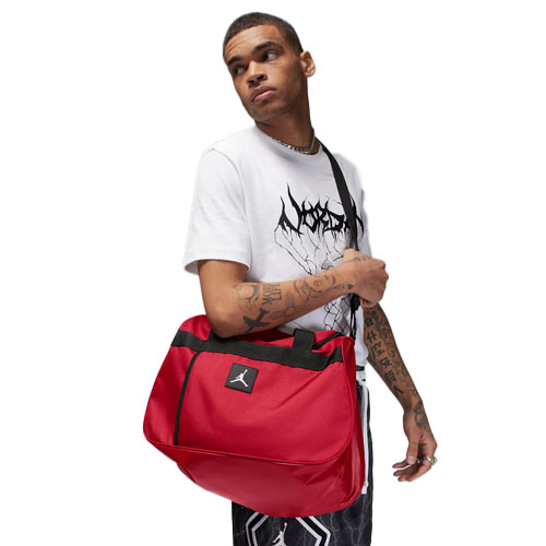 A person carries a red Jordan messenger bag featuring the Jumpman logo, wearing a white graphic t-shirt and patterned shorts.