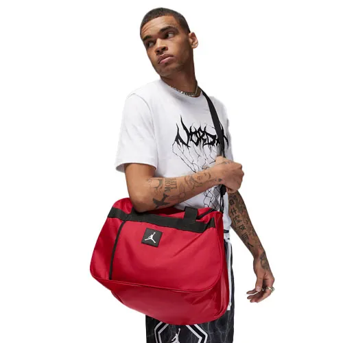 A person carries a red Jordan messenger bag featuring the Jumpman logo, wearing a white graphic t-shirt and patterned shorts.
