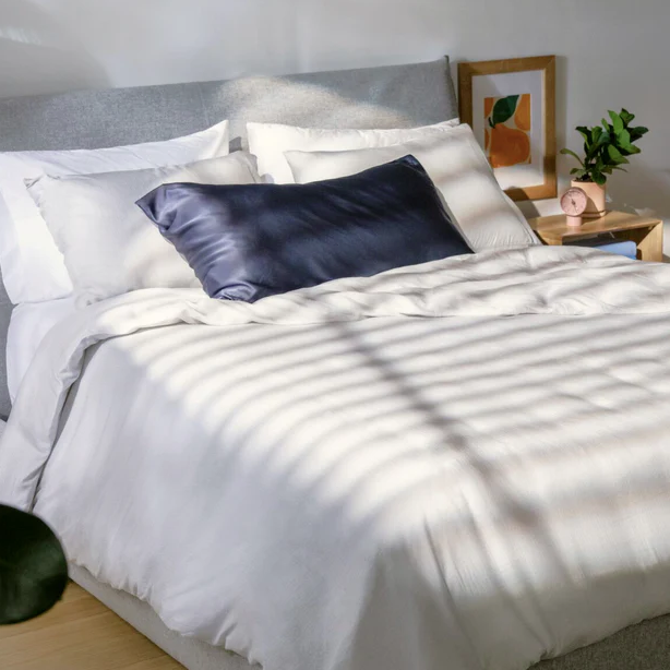 A neatly made bed with white bedding and a navy pillow is next to a wooden nightstand featuring a small plant, a pink alarm clock, and framed art on the wall.