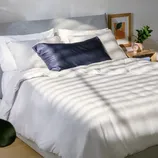 A neatly made bed with white bedding and a navy pillow is next to a wooden nightstand featuring a small plant, a pink alarm clock, and framed art on the wall.