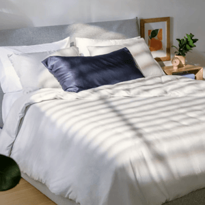 A neatly made bed with white bedding and a navy pillow is next to a wooden nightstand featuring a small plant, a pink alarm clock, and framed art on the wall.