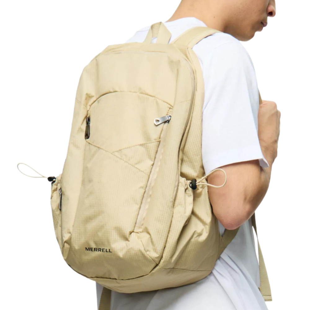 A beige Merrell backpack worn by a person in a white t-shirt, featuring multiple zippered compartments and adjustable straps.