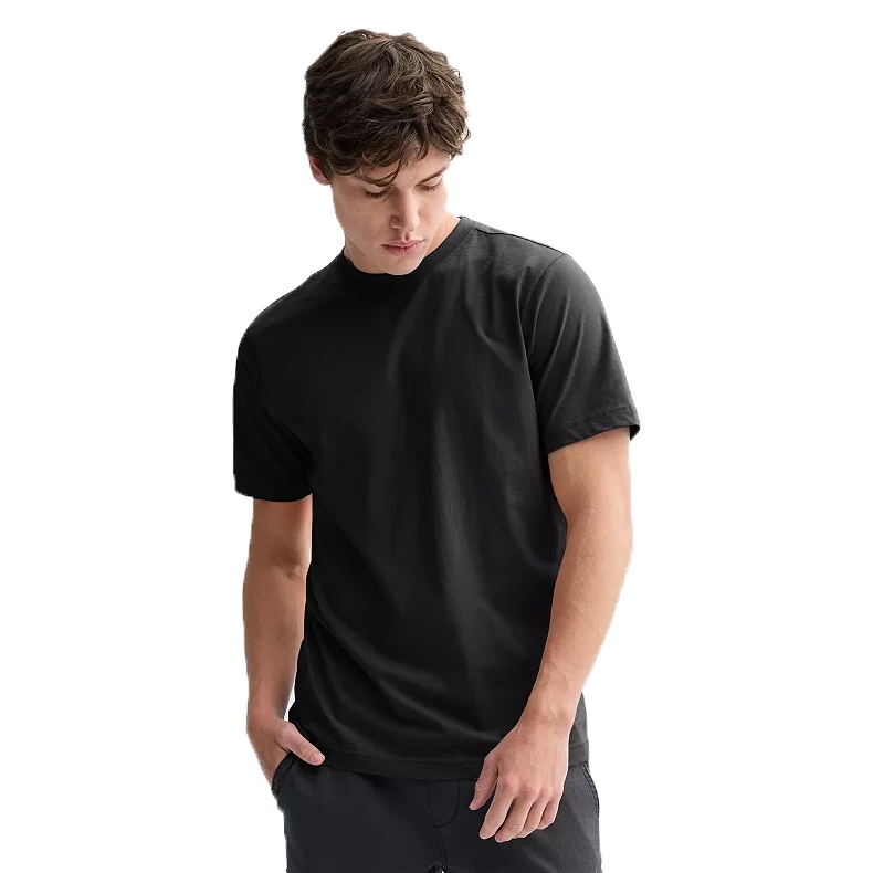 A man wearing a plain black T-shirt with short sleeves. He is casually posed with one hand in a trouser pocket, looking downward.